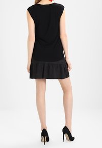 Kvinna i en ärmlös svart klänning med volangdetaljer, röda örhängen och svarta högklackade skor, stående med ryggen mot kameran.