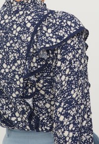 Námořnická modrá květinová blůza s bílým vzorem, volánkovým výstřihem a texturovanou tkaninou. Obsahuje malé květiny a detailní prošívání podél okrajů.