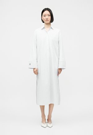 Femme debout portant une longue robe chemise blanche à fines rayures, avec des poignets larges, une poche poitrine rayée et des chaussures blanches à bouts pointus.