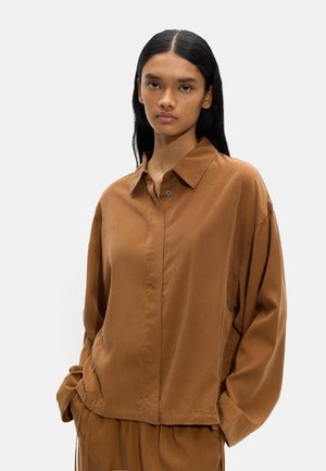 Młoda kobieta z długimi czarnymi włosami, ubrana w luźną brązową koszulę i pasujące spodnie, stojąca na tle jednolitego jasnego tła.