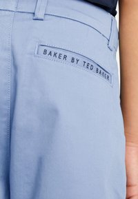 Hellblaue Hose aus glattem Stoff, mit taillierter Passform und einem eingenähten Etikett mit der Aufschrift "BAKER BY TED BAKER" auf der Hintertasche.