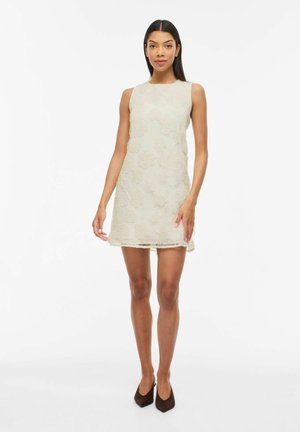 Kvinde står iført en ærmeløs mini-kjole i cremefarvet blonder og sorte spidse hæle på en hvid baggrund.