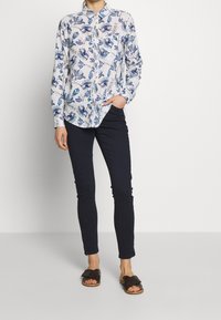 Mujer vestida con una camisa blanca de botones con un patrón floral azul, pantalones negros ajustados y sandalias deslizantes negras, contra un fondo liso.