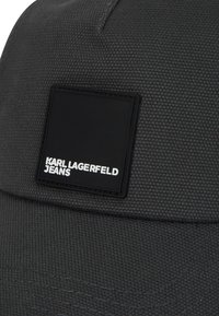 Căciulă de culoare gri închis, realizată dintr-un material texturat, având o etichetă pătrată neagră cu textul alb "KARL LAGERFELD JEANS". Vizor curbat și cusături îngrijite.