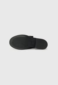 Black rubber shoe sole with textured grip pattern; features a small gold buckle accent and a rounded heel.