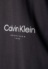 Musta kangas, jossa valkoisella painettuna teksti "Calvin Klein" ja "Perustettu 1968" yksinkertaisella, modernilla fontsilla.