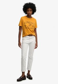 Geel katoenen T-shirt met "SUPERDRY" grafisch ontwerp, gecombineerd met witte jeans en donkere loafers. Eenvoudig design en klassieke pasvorm.