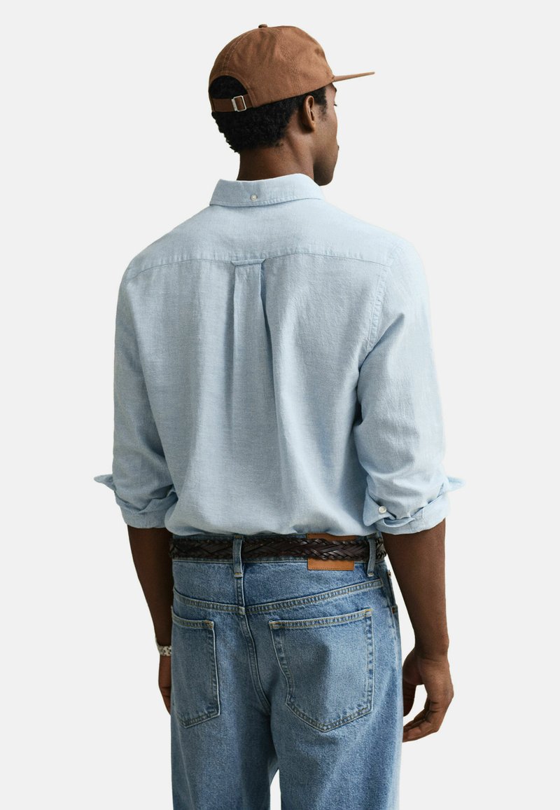 Mand set bagfra iført lyseblå langærmet skjorte med op rullede ærmer, blå jeans, brun bælte, brun kasket og sølvfarvet armbåndsur.