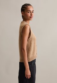 Beige knitted sleeveless sweater with a textured finish and ribbed hem. Paired with black pants, it features a relaxed and loose fit.