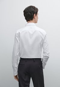 Weißes Hemd mit klassischem Kragen, glatter Textur, langen Ärmeln und dunklen, engen Hosen mit Gesäßtaschen, von hinten gezeigt.