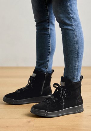 Zwarte suède hoge laarzen met ronde neus, vetersluiting, zijrits en rubberen zool. Gecombineerd met strakke blauwe jeans.