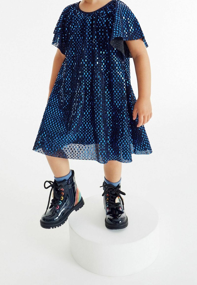 Enfant debout sur une plateforme ronde blanche portant une robe bleue scintillante et des bottes en cuir verni noir avec des détails arc-en-ciel.
