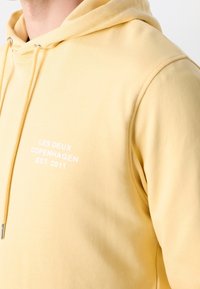 Homme portant un hoodie jaune pâle avec le texte blanc "LES DEUX COPENHAGEN EST. 2011" sur la poitrine et des cordons de serrage avec des embouts en argent.