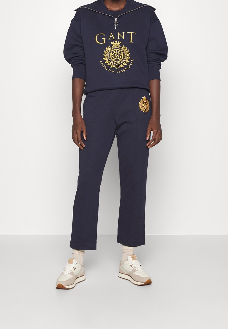 Personne portant un sweat-shirt GANT bleu marine et un pantalon assorti avec un logo jaune et des baskets beiges, debout devant un fond blanc.
