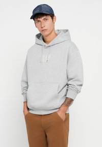 Sudadera gris claro con bolsillo frontal y capucha con cordones; presenta un pequeño logo de GAP. Se lleva con pantalones marrones y una gorra texturizada azul marino.