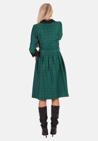 Grünes Karokleid mit tailliertem Oberteil und plissiertem Rock. Mit schwarzem Kragen und langen Ärmeln, kombiniert mit kniehohen schwarzen Stiefeln.