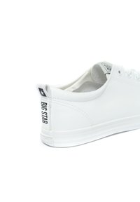 Bílé kožené tenisky s texturovaným povrchem, s černým logem na patě a plochou bílou gumovou podrážkou. Jednoduchý design pro provlékání šňůrek.