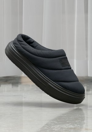 Black quilted slip-on shoe with a cushioned collar and textured outsole. Features a logo patch on the side and a rounded toe design.