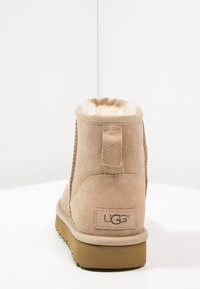 Bota de ante color tierra con un suave forro de borrego, de altura baja en el tobillo y suela de goma texturizada. Presenta una pequeña pestaña en la parte trasera y un detalle del logo.