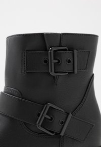 Svarta läderankelstövlar med två justerbara svarta spännen, slät yta och rundad topp. Sytt accentuerade detaljer är synliga.