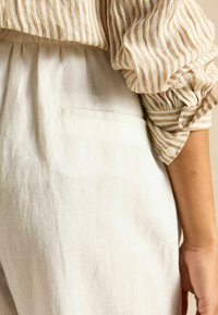 Prúžkovaný béžovo-biely ľanový top s nariasenými rukávmi, skombinovaný s ľahkými ľanovými nohavicami svetlej farby s zadným vreckom.