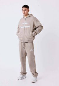 Beige hoodie en sweatpants set met een ritssluiting, voorzakken en wit "PXP" logo. Strak ontwerp met een zachte textuur. Witte sneakers.
