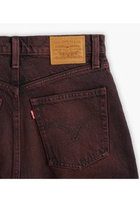 Jeans de mezclilla rojo oscuro con un parche de cuero etiquetado "Levi Strauss & Co." Presenta un bolsillo trasero con la icónica etiqueta de Levi's y detalles de costura.