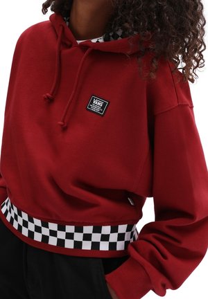 Person iført en rød Vans hættetrøje med en sort-hvid ternet kant og en lille Vans logo-patch på brystet.