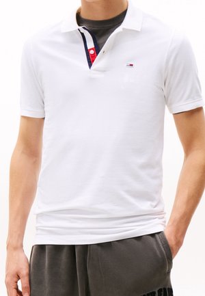 Homme portant un polo blanc avec un petit logo sur la poitrine et une patte de boutonnage rayée sur un T-shirt foncé, associé à un pantalon sombre.