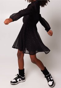 Černé, průhledné šaty s dlouhým rukávem, s nařaseným pasem a třpytivým tečkovaným detailem. Nosené s černobílými teniskami a černými ponožkami.