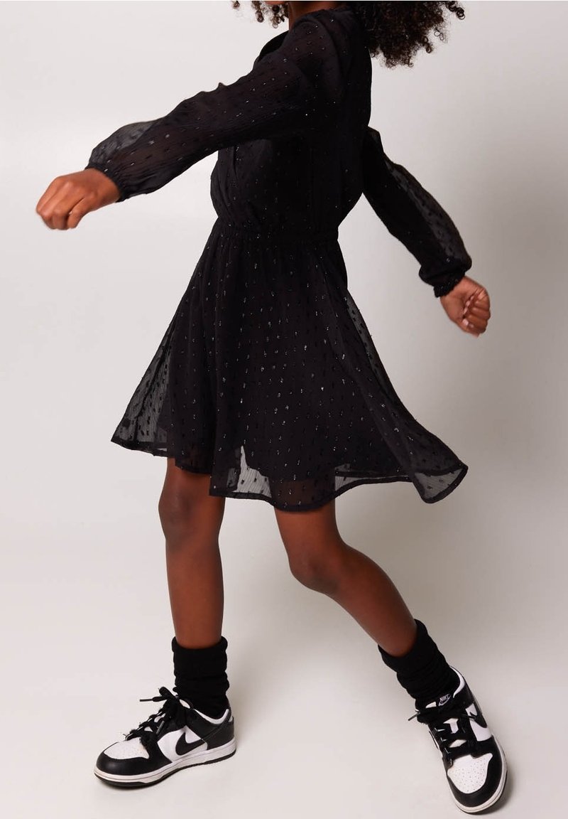 Černé, průhledné šaty s dlouhým rukávem, s nařaseným pasem a třpytivým tečkovaným detailem. Nosené s černobílými teniskami a černými ponožkami.