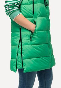 Grüne Steppweste mit Reißverschlusstaschen, die ein gepolstertes Design und schwarze Akzentreißverschlüsse aufweist. Das Material wirkt glänzend und glatt.