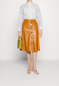 Blouse brodée bleu clair avec des découpes, associée à une jupe en cuir marron avec des boutons. Tenant un petit sac à main vert.