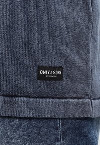 Blaues Strick-Sweatshirt mit geripptem Saum, versehen mit einem schwarzen Logostreifen mit der Aufschrift "ONLY & SONS" nahe dem unteren Rand. Strukturierter Stoff, lässiges Design.