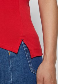 Top de algodón rojo con un patrón texturizado y aberturas laterales. Combinado con denim azul, mostrando detalles de costura en los vaqueros.
