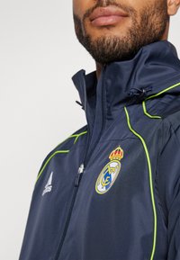 Mornarska jakna s kapuljačom s zelenim detaljima, zatvaranje na zip, visoki ovratnik i logotip Real Madrida na lijevoj strani prsnog koša; izrađena od glatkog, laganog materijala.