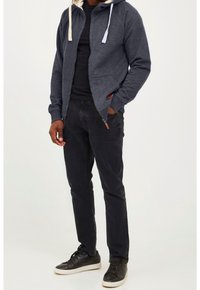 Sudadera con cremallera de color gris oscuro con una capucha interior de color gris claro en contraste, combinada con jeans negros y zapatillas negras. Cuenta con bolsillos laterales y cordones ajustables.