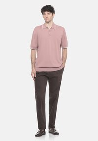 Giovane in piedi con le mani in tasca, indossa una polo a maniche corte color malva, pantaloni marroni scuri e mocassini scuri.