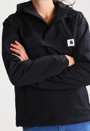 Persoon met gekruiste armen, gekleed in een zwarte Carhartt capuchonjas en een blauwe spijkerbroek tegen een effen achtergrond.