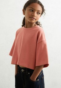 Fiatal lány, göndör hajjal, lazán oversized, rózsaszínű cropped pólóban és sötét farmernadrágban, kezei a zsebében, oldalra néz egy sík háttér előtt.