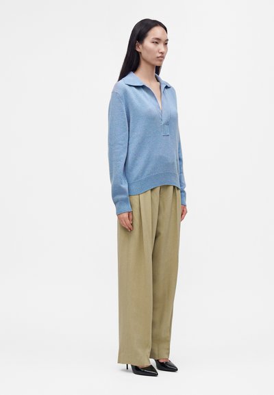 Femme debout portant un pull bleu clair à col, un pantalon beige large et des chaussures à talons hauts pointues noires sur un fond blanc uni.