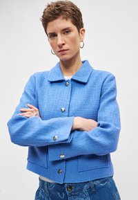 Κοντό μπλε σακάκι από τσόχινο ύφασμα με γιακά, διακοσμημένο με ασημένια κουμπιά και δύο μπροστινές τσέπες. Η υφή είναι υφασμένη και ελαφρώς τραχεία.