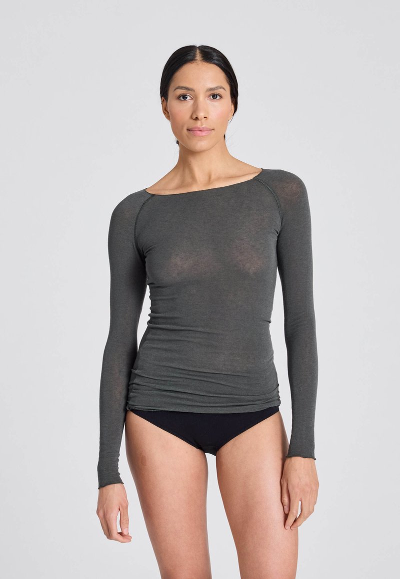 Femme aux cheveux foncés tirés en arrière, portant un haut gris foncé ajusté à manches longues et des sous-vêtements noirs, debout devant un fond clair uni.