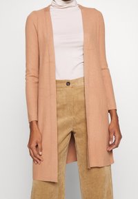 Pfirsichfarbene gerippte Strickjacke mit langen Ärmeln, offenes Vorderdesign, weicher Stoff, getragen über einem hellen Rollkragenpullover und beigen Cordhosen.