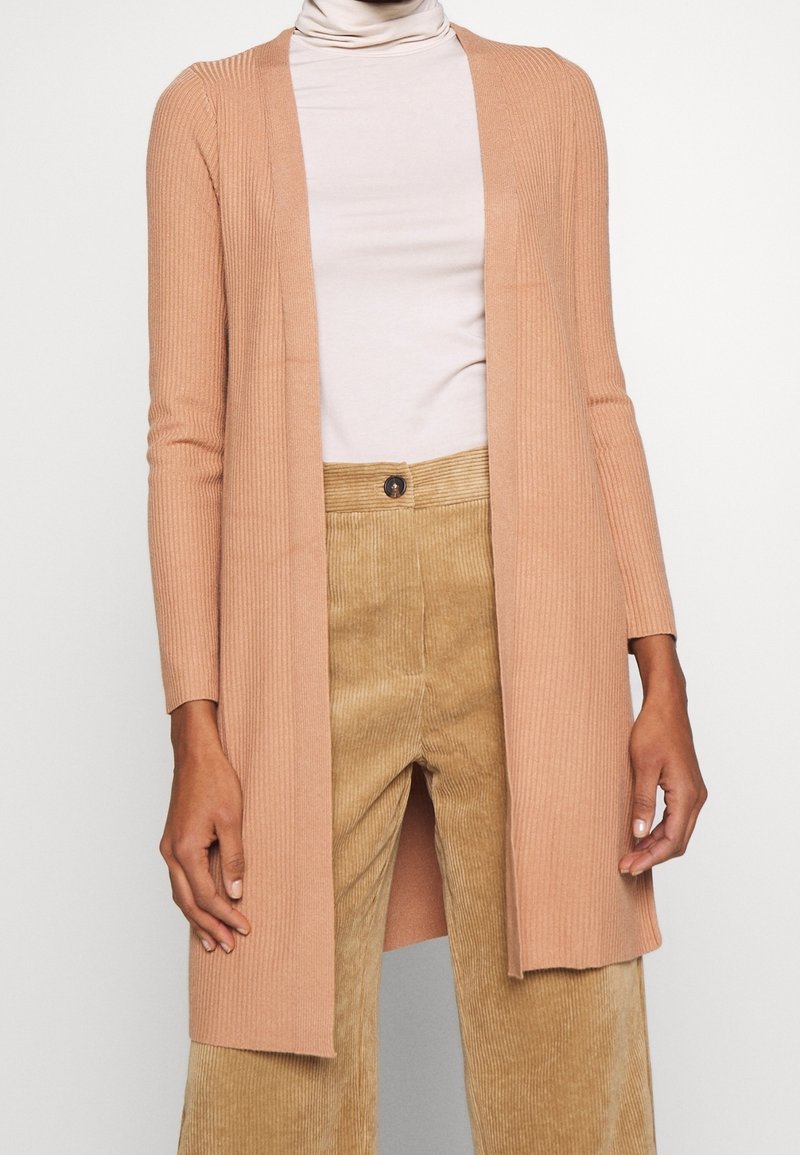 Pfirsichfarbene gerippte Strickjacke mit langen Ärmeln, offenes Vorderdesign, weicher Stoff, getragen über einem hellen Rollkragenpullover und beigen Cordhosen.