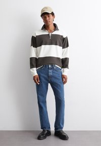 Hosszú ujjú, cipzáras pulóver sötétbarna és fehér csíkokkal, közepesen mosott kék farmernadrággal és fekete cipővel párosítva. Bézs sapka a fején.