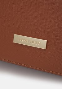 Brązowa torebka o teksturowanej powierzchni z złotą metalową płytką z wyrytą nazwą "ANNA FIELD" i widocznymi szwami na brzegach.