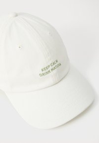 Boné branco com tecido texturizado e orifícios de ventilação costurados. Texto verde bordado que diz "MANTENHA A CALMA E BEBA MATCHA." Aba curvada.