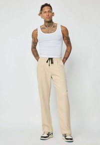 Pantalons de survêtement beiges avec taille à cordon de serrage et poches latérales, associés à un débardeur blanc et des baskets noir et blanc.