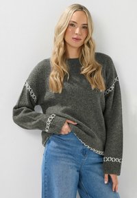 Grijze gebreide trui met een relaxte pasvorm, voorzien van witte geborduurde accenten op de mouwen en de zoom. Gecombineerd met blauwe denim jeans.